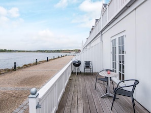 Een houten terras met een barbecue, twee stoelen en een tafel, met uitzicht op een waterpartij.