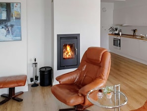 Een moderne woonkamer met een leren fauteuil, een glazen salontafel, een open haard en een keuken op de achtergrond.