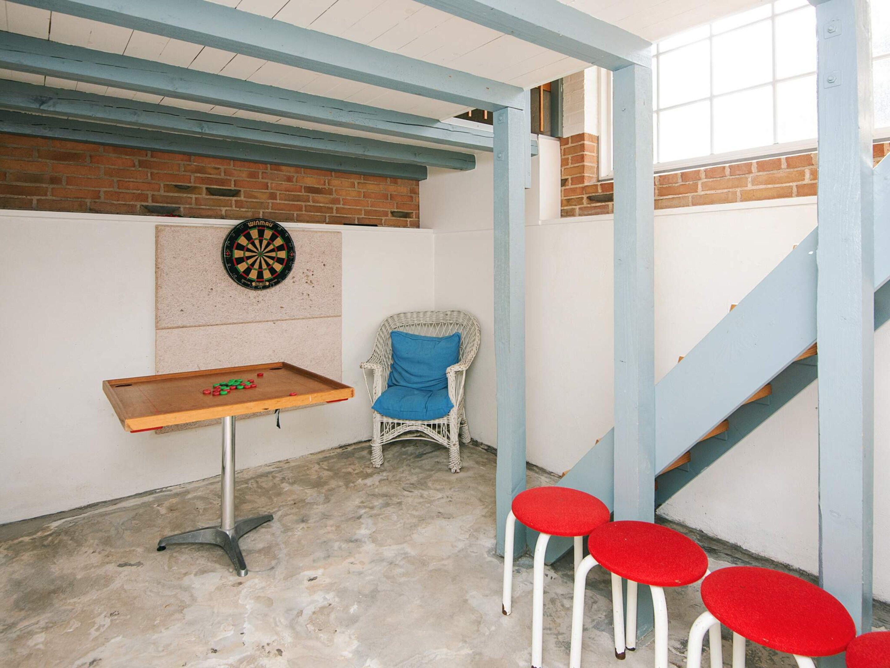 A room with a dartboard, a small table with playing cards, a wicker chair, and red stools.