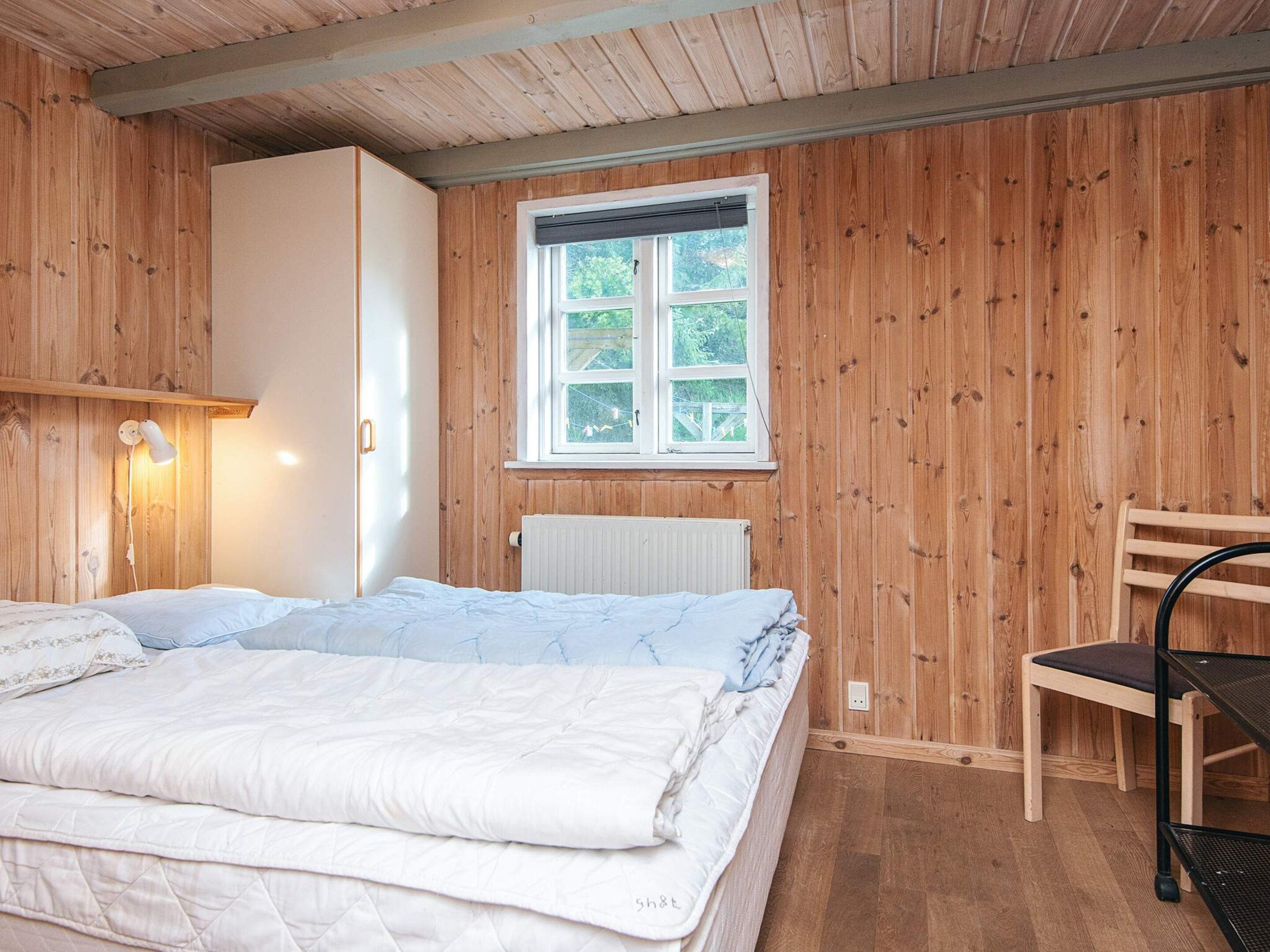 A bedroom with a bed, a chair, a window, and wooden walls.