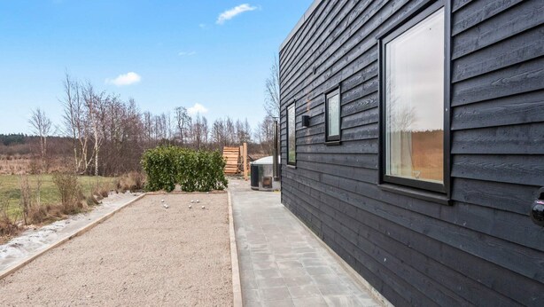 A modern building with a dark exterior, a paved pathway, and a small garden area with a bush and gravel. - Holiday Home in Glesborg (Glesborg)