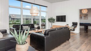 A modern living room with a large window, a black leather sofa, a wooden coffee table, and a pool table. - Holiday Home in Glesborg (Glesborg)