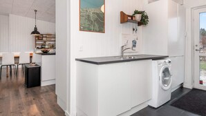A modern kitchen with a built-in washing machine, a dining area with a wooden table and chairs, and a wall-mounted shelf with a plant. - Holiday Home in Glesborg (Glesborg)