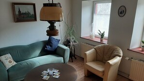 A living room with a green sofa, a beige armchair, a round table with playing cards, a lamp, a potted plant, and a window with a view of a building. - Beautiful Holiday Home With Terrace (Nattenheim)
