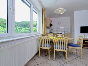 A dining area with a table and chairs, a kitchen with a clock, and a window with a view of greenery.