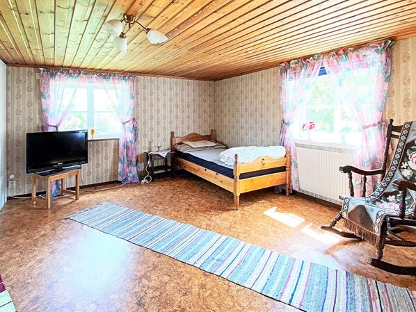 A bedroom with a wooden ceiling, a bed, a television on a stand, a rocking chair, and a window with curtains.