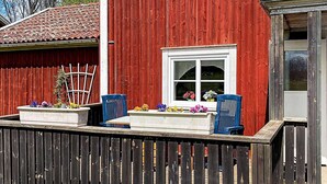 A red wooden house with a white window and a small porch area with two blue chairs and flower planters. - 7 Person Holiday Home in Almedboda (Älmeboda)