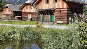 Une cabane en bois dotée d’un porche et d’un petit banc, entourée d’un étang et de hautes herbes.