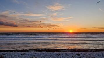 Pôr do sol sobre o oceano, com uma praia de pedrinhas em primeiro plano.