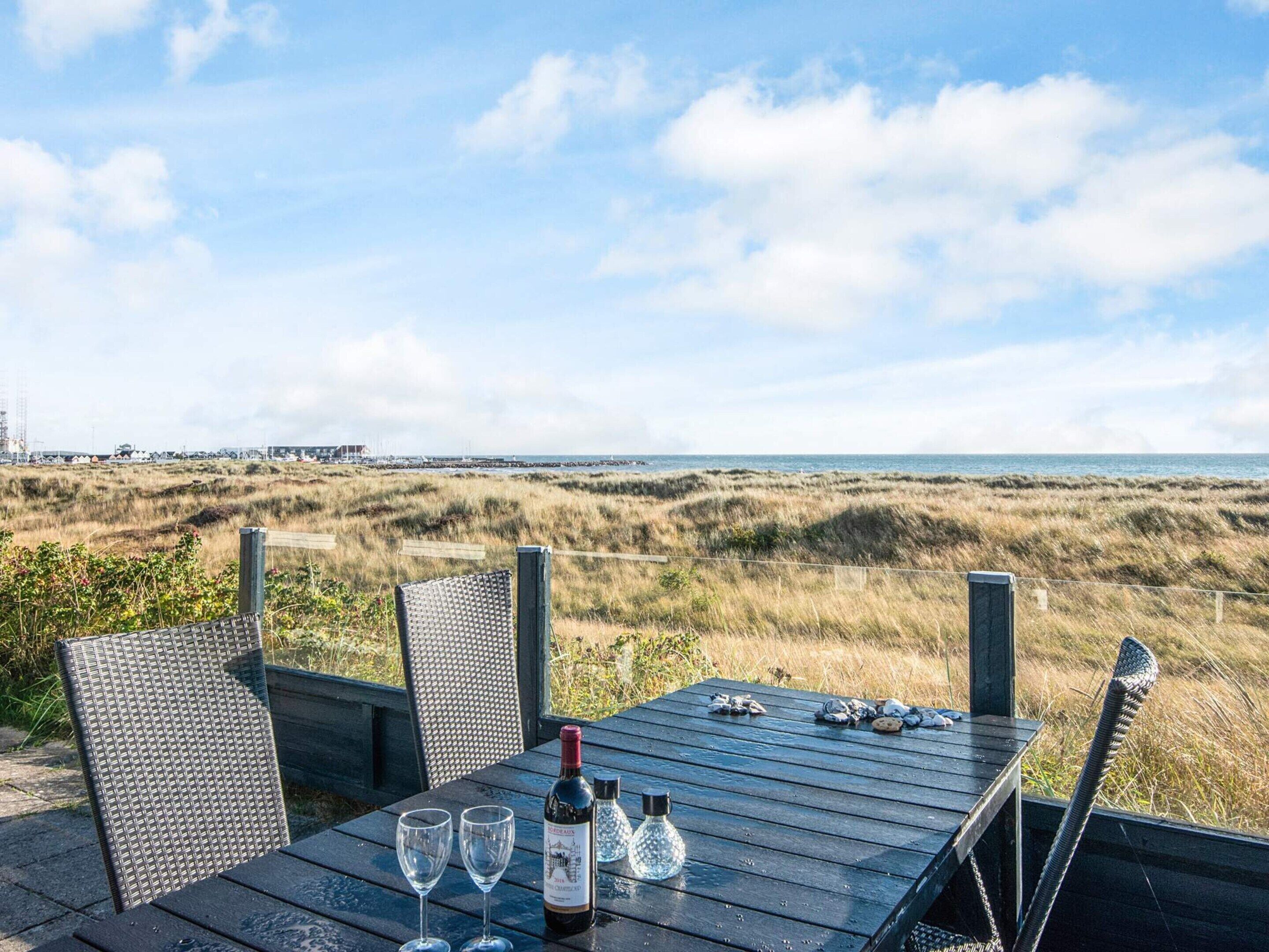 Uma mesa com duas taças de vinho e uma garrafa, com vista para a costa, dunas e um cais distante.