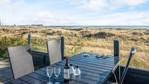 A table with two wine glasses and a bottle, set against a coastal view with dunes and a distant pier.