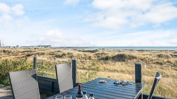Uma mesa com duas taças de vinho e uma garrafa, com vista para a costa, dunas e um cais distante.
