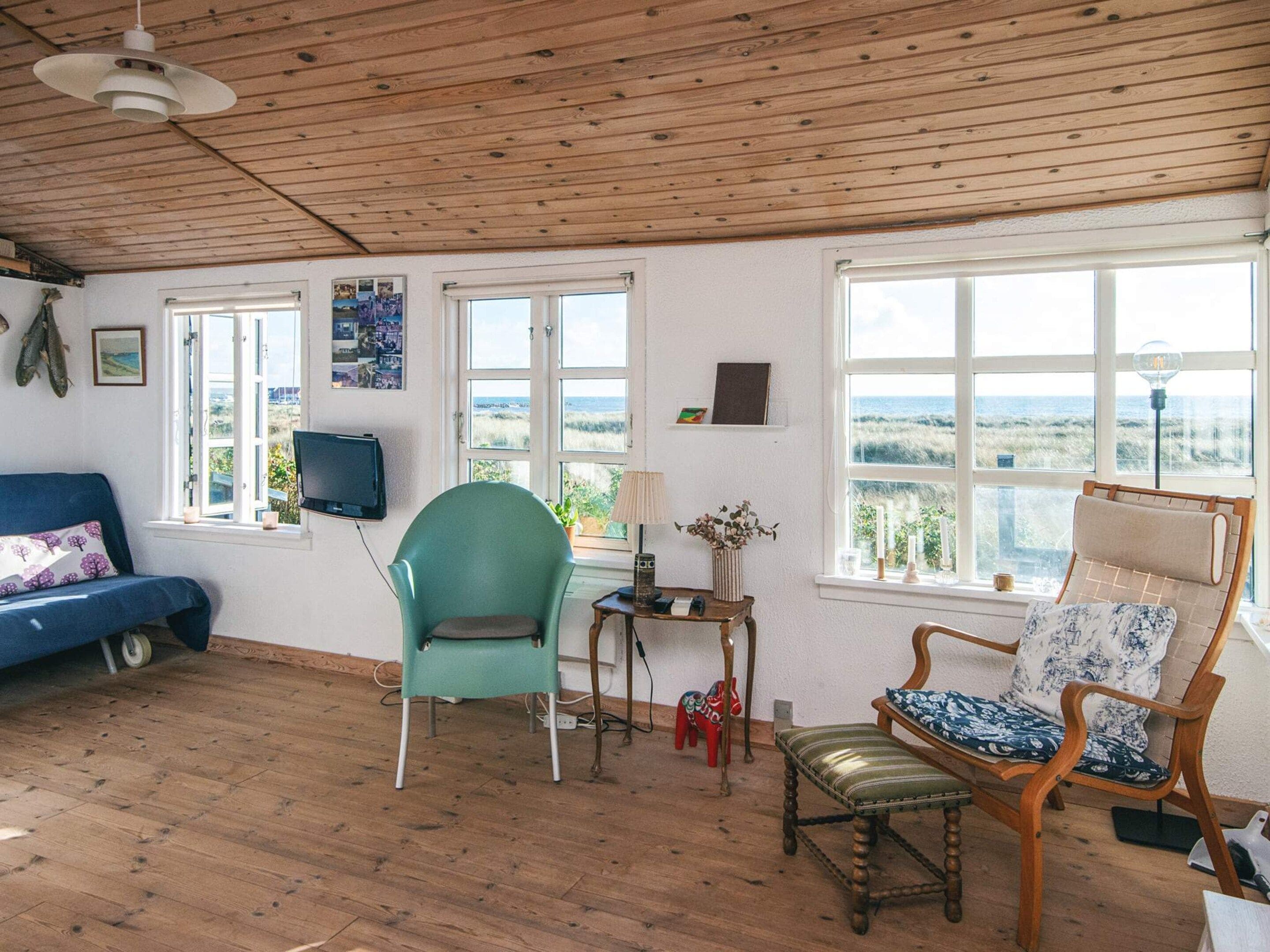 Una habitación con techo de madera, un sofá azul, una silla verde, un banco de madera y ventanas amplias que dan a vista al mar.