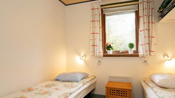Une petite pièce agréable et chaleureuse, avec deux lits simples, une table de chevet en bois, une fenêtre avec des rideaux et des lampes fixées au mur.