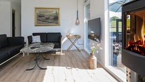 A modern living room with a black sofa, a round coffee table, a painting on the wall, a fireplace, and a TV mounted on the wall. - Holiday Home in Idestrup (Idestrup)