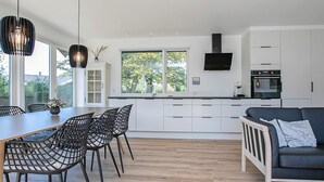 A modern kitchen with white cabinets, a dining area with black chairs, and a grey sofa. - Holiday Home in Idestrup (Idestrup)