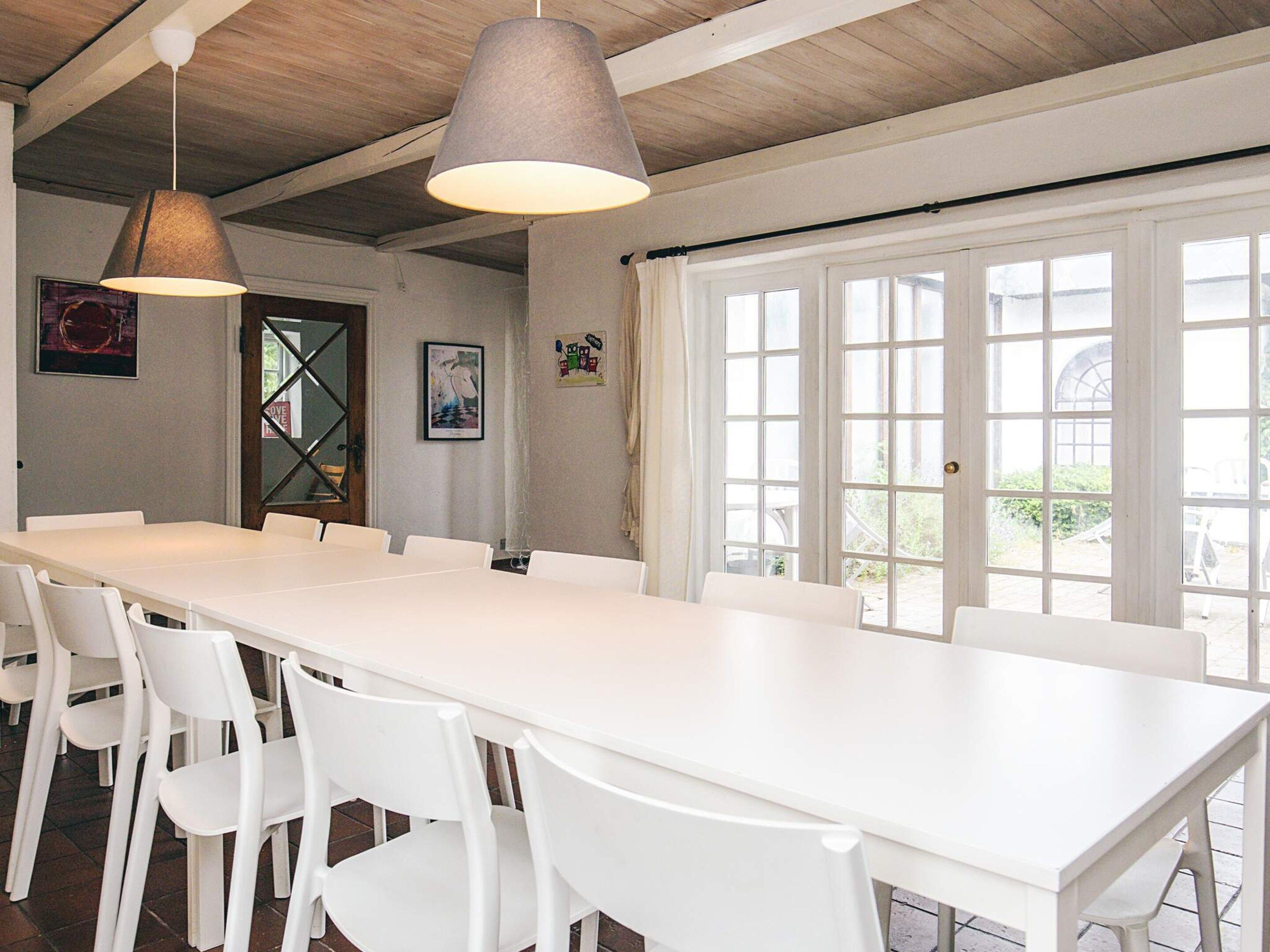 A long white table with chairs in a room with wooden beams on the ceiling, large windows, and a door with glass panels.