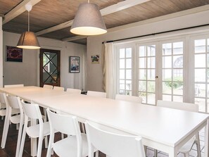 A long white table with chairs in a room with wooden beams on the ceiling, large windows, and a door with glass panels. - Holiday Home in VÃ¦ggerlÃ¸se (Vaeggerlose)