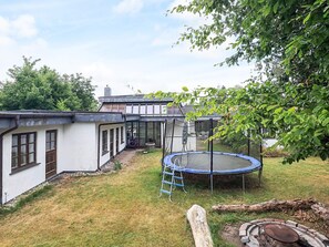 Un jardin à l’arrière d’une maison, aménagé avec un trampoline, une terrasse en bois et un foyer en briques.