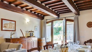 Un coin repas avec une table mise pour le repas, des poutres en bois au plafond, une vue sur la verdure à l’extérieur et un meuble contenant des bouteilles.