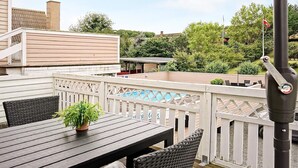 Une terrasse en bois avec une table et des chaises, entourée d’une clôture blanche, donnant sur une piscine et un espace verdoyant.