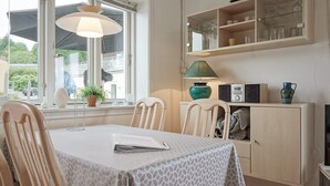 Un coin repas avec une table recouverte d’une nappe à motifs, des chaises en bois, une lampe et un buffet contenant de la verrerie et une radio.