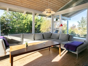 Una sala moderna con un sofá seccional, una mesa de centro de madera y grandes ventanales que ofrecen vistas a árboles y una casa.