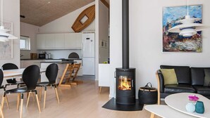 Ett modernt vardagsrum med en central vedeldad kamin, en matplats med stolar och ett bord, och ett kök i bakgrunden.