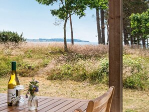 Une table en bois, sur laquelle sont posés une bouteille de vin, un verre et un vase rempli de fleurs, le tout sur fond d’un champ verdoyant et d’arbres.