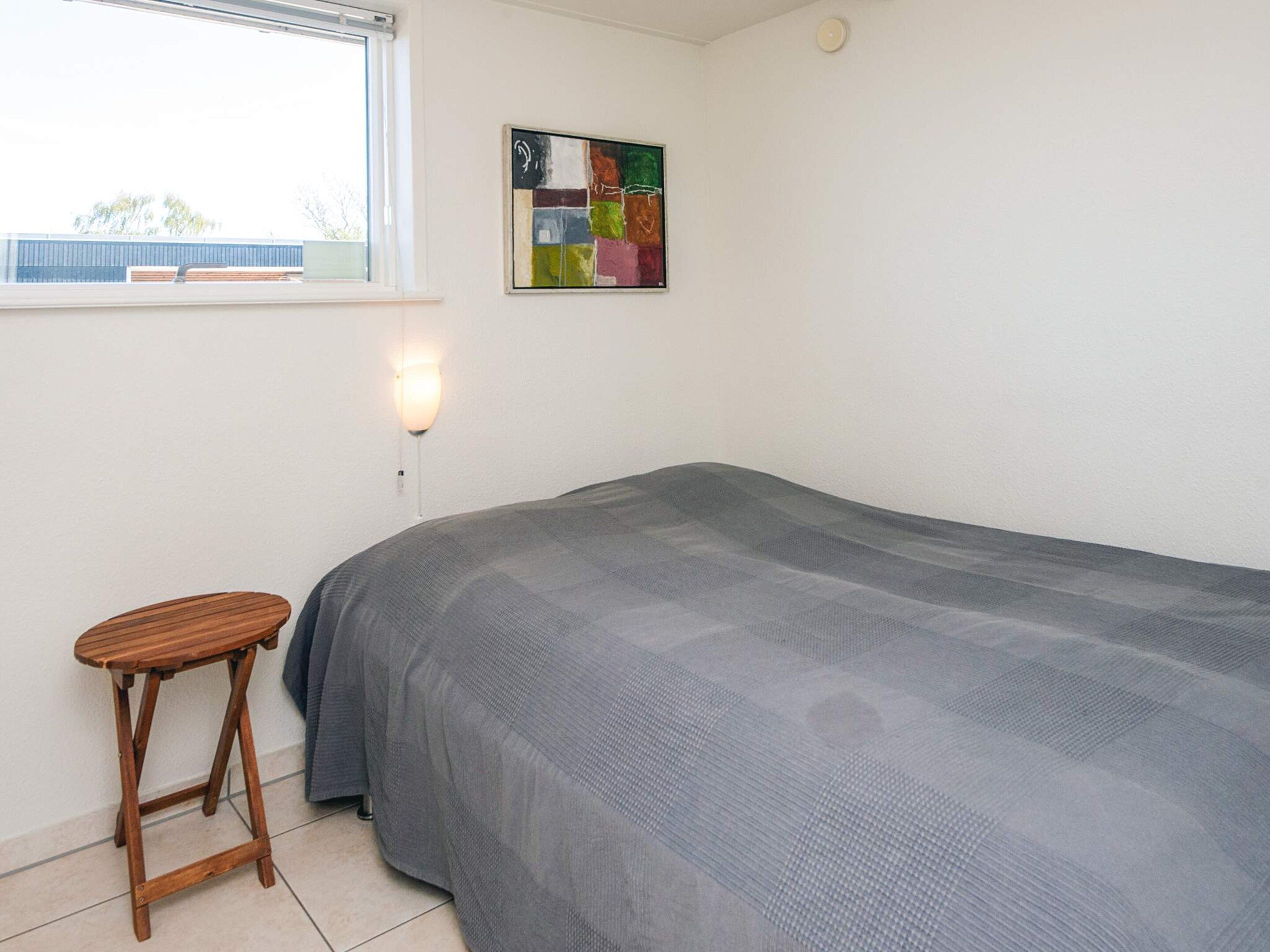 A single bed with a grey blanket, a wooden stool, a wall-mounted light, and a colorful abstract painting.