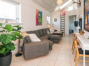A modern living room with a sectional sofa, a dining area with wooden chairs, a large potted plant, and a painting on the wall.