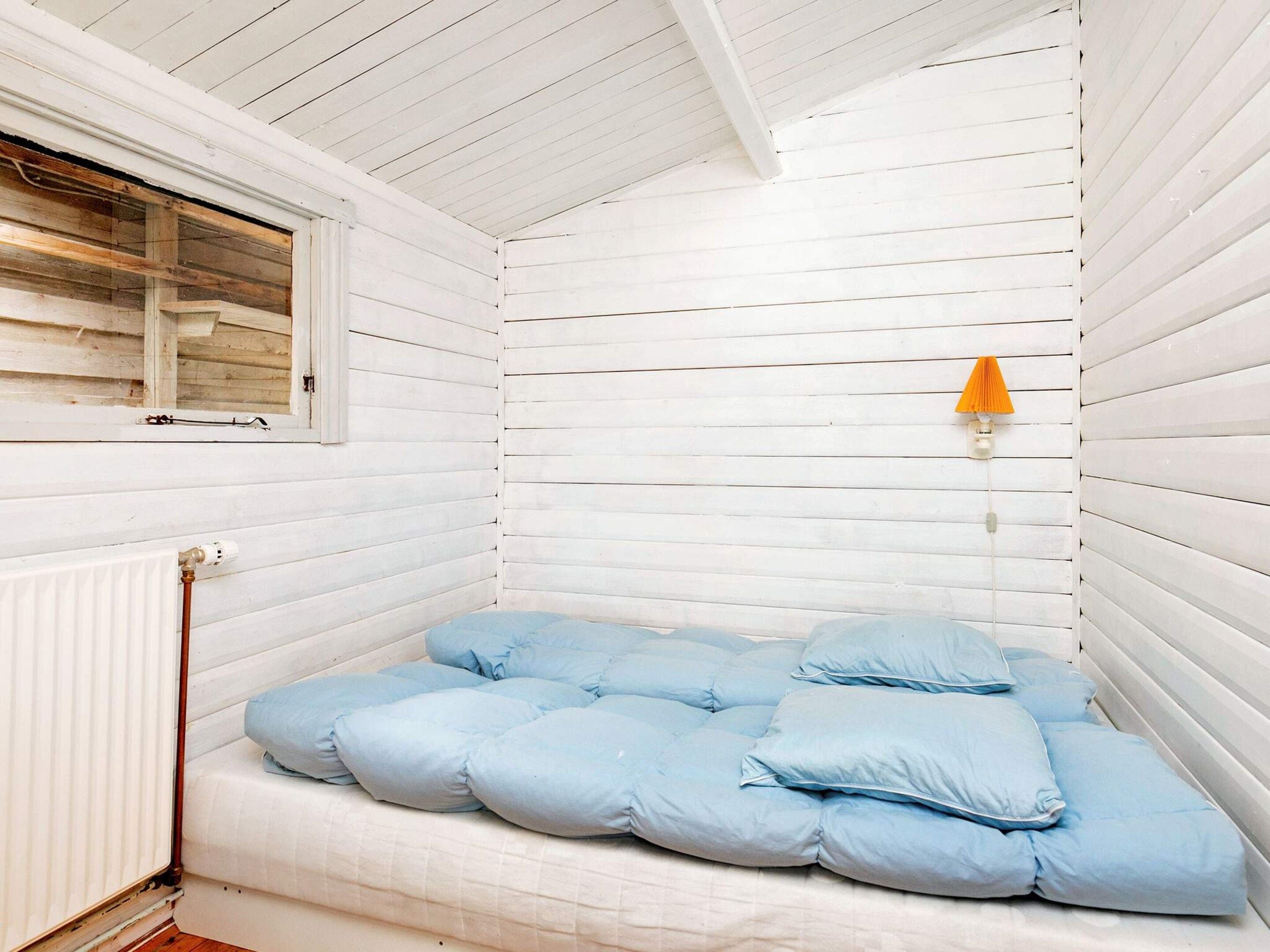 A small, white-paneled room with a bed, a window, and a wall-mounted lamp.