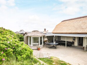 A covered outdoor seating area with a thatched roof, surrounded by lush greenery and a garden. - 16 Person Holiday Home in Ulfborg (Ulfborg)
