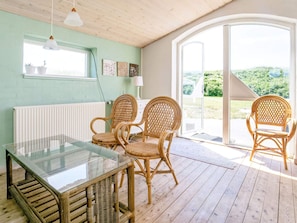Quarto com mesa de vidro, cadeiras de vime, radiador branco e vista para área externa através de uma porta de vidro.