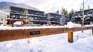 A snow-covered wooden fence with a 'VISITOR' sign, modern buildings in the background, and a mountainous landscape. - Cozy and Modern Getaway (Revelstoke)