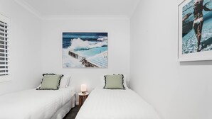 A bedroom with two beds, a nightstand with a lamp, a large ocean view painting, and a smaller framed picture. - Coogee by the Coast  (Coogee)