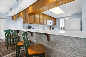 Eine moderne Küche mit Holzschränken, einer weißen Arbeitsplatte und einer Granitspritzschutzleiste. Zwei grüne Stühle und ein Holztisch sind ebenfalls vorhanden.