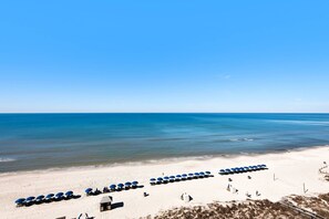 Een strand met rijen blauwe parasols en ligbedden, een wachthuisje en een helder uitzicht op de oceaan.