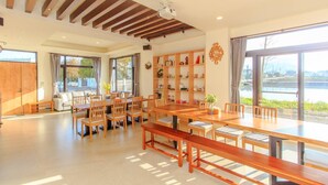 A spacious dining area with wooden tables and chairs, a bookshelf with various items, and large windows offering a view of a body of water. - Classic Villa B&B (Dongshan)