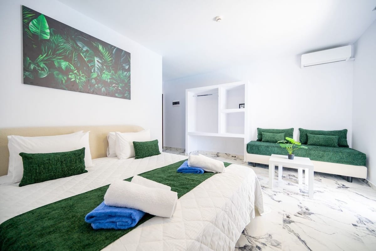 Una habitación de hotel moderna con dos camas, ropa de cama verde y blanca, una planta verde sobre una mesita de noche y un cuadro grande con estampado de hojas en la pared.