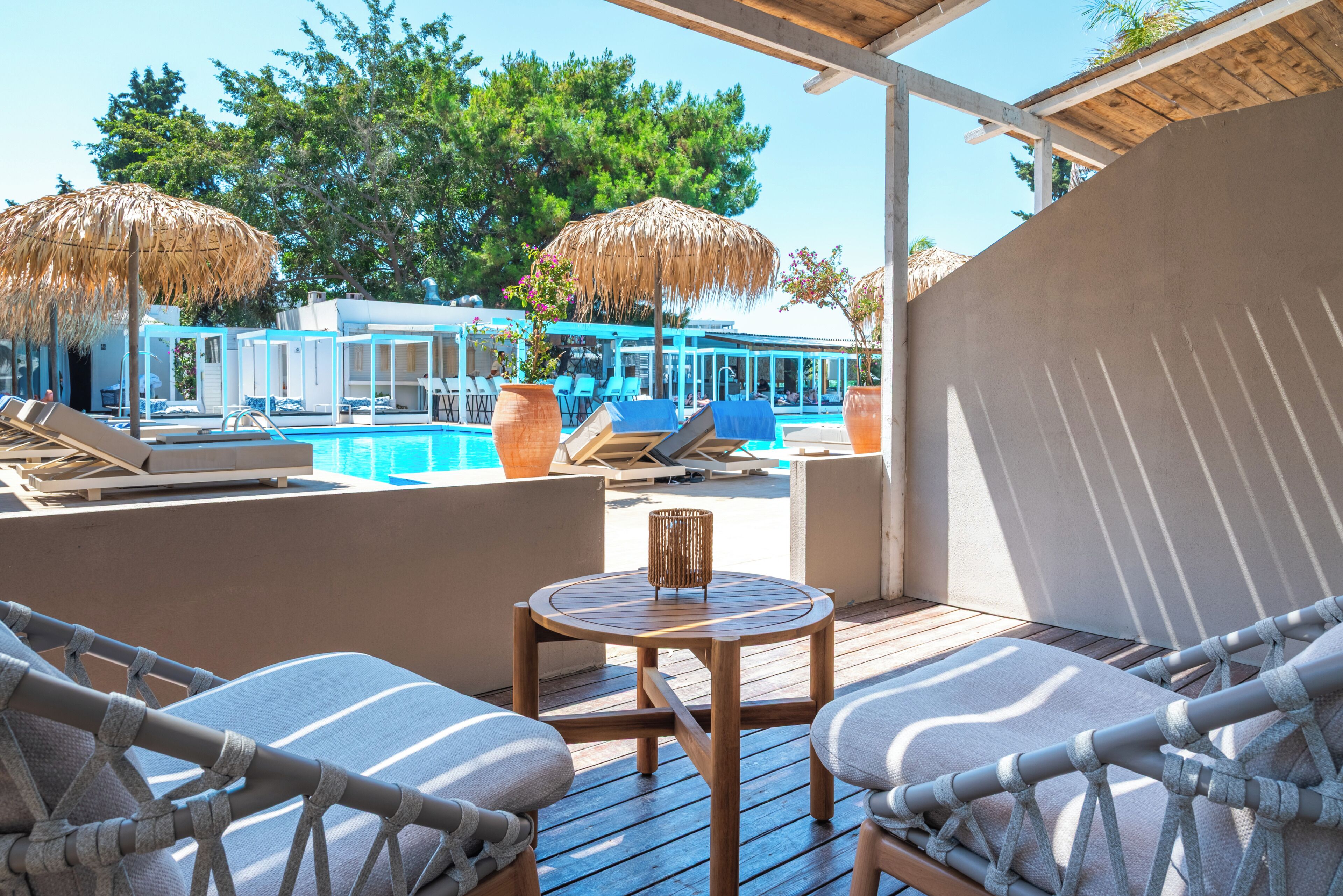 Zona junto a la piscina con sillones de descanso, una mesa de madera y vistas a la piscina y árboles.