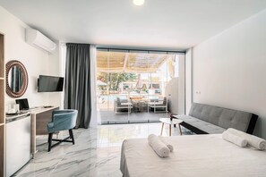 A modern hotel room with a bed, desk, TV, and a view of an outdoor pool area. - Bamboo Suites Adults Only (Rhodes)