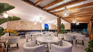 Un restaurante con mesas redondas y sillas blancas, paredes de piedra y lámparas colgantes.