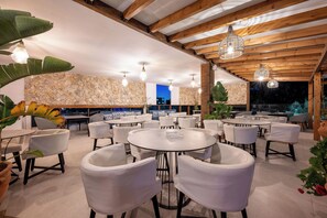 A restaurant with round tables and white chairs, stone walls, and hanging light fixtures. - Bamboo Suites Adults Only (Rhodes)