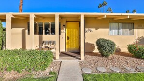 A single-story building with a yellow door, a bench outside, and a small garden with pebbles and plants. - Mid-Century Beauty! Community Pool! Hiking! Mountain Views! Walk to El Paseo! (Palm Desert)