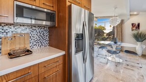 A modern kitchen with wooden cabinets, a stainless steel refrigerator, and a microwave. There is a dining area with a table and chairs visible. - Mid-Century Beauty! Community Pool! Hiking! Mountain Views! Walk to El Paseo! (Palm Desert)