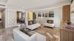 A modern living room with two sofas, a coffee table, and a rug. There are windows with a view of greenery outside. - Mid-Century Beauty! Community Pool! Hiking! Mountain Views! Walk to El Paseo! (Palm Desert)