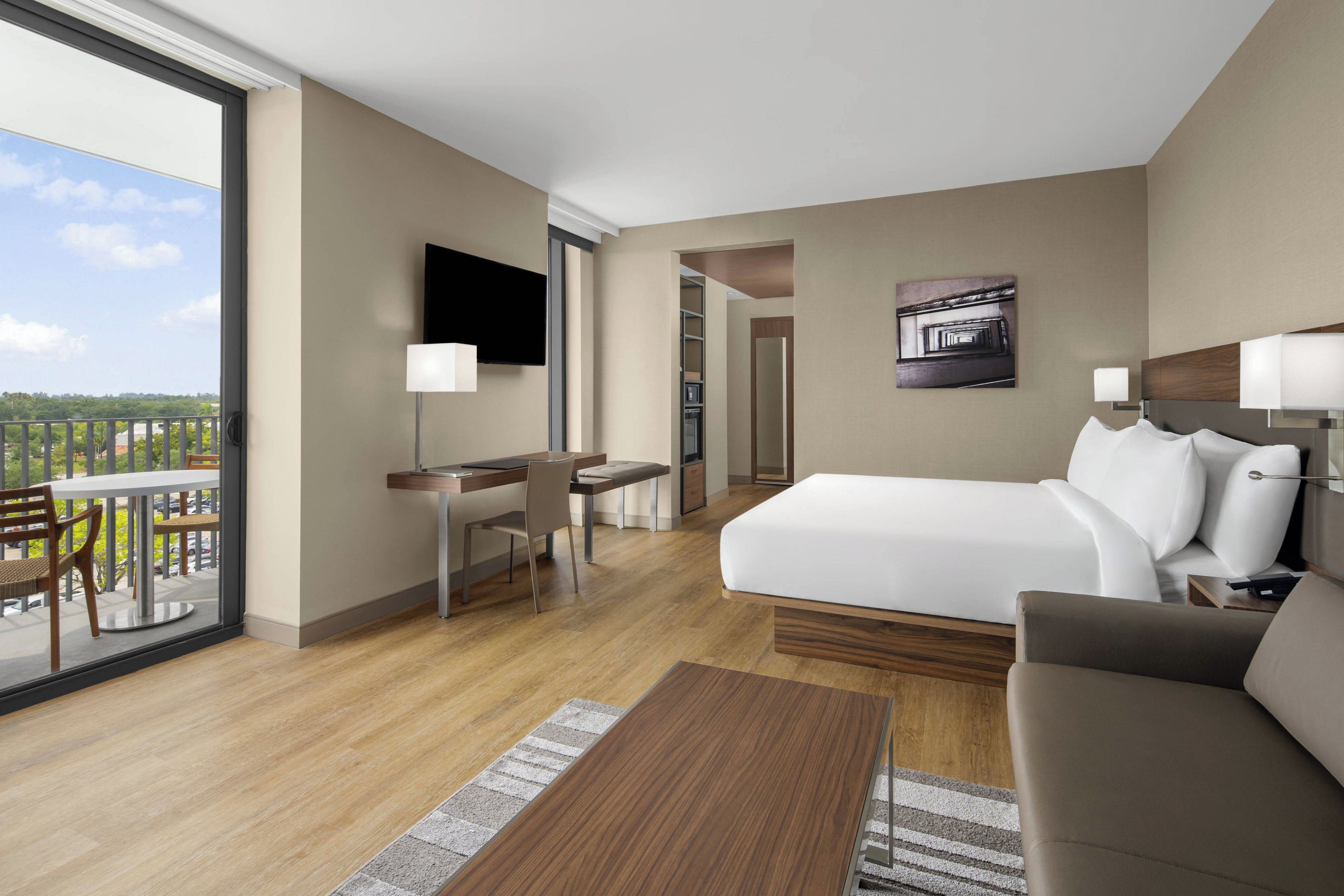 A hotel room with a bed, a desk, a chair, a TV, and a balcony with a view of the city.
