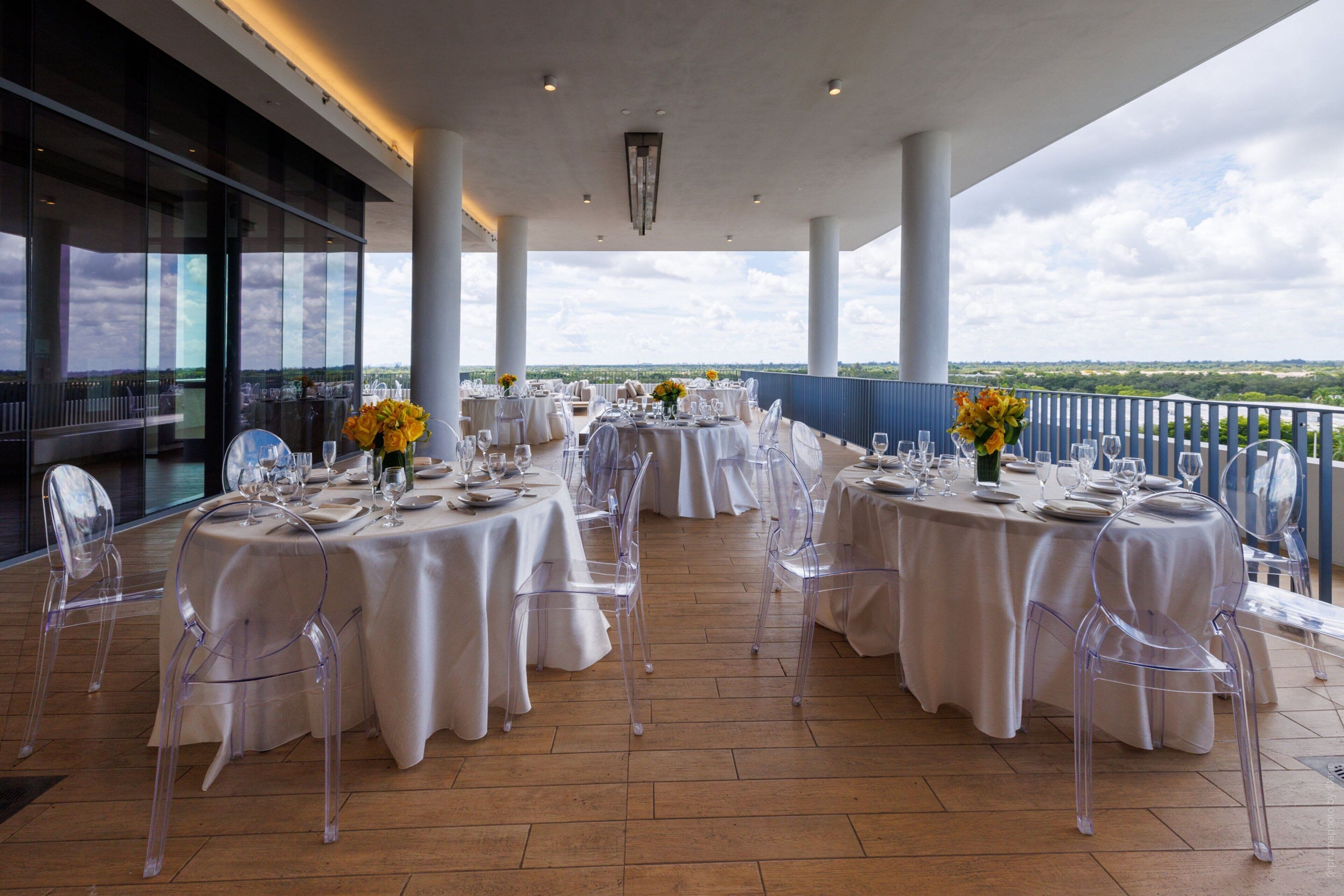 Un área de comedor al aire libre espaciosa con varias mesas redondas dispuestas para una comida, sillas traslúcidas y vistas al paisaje.