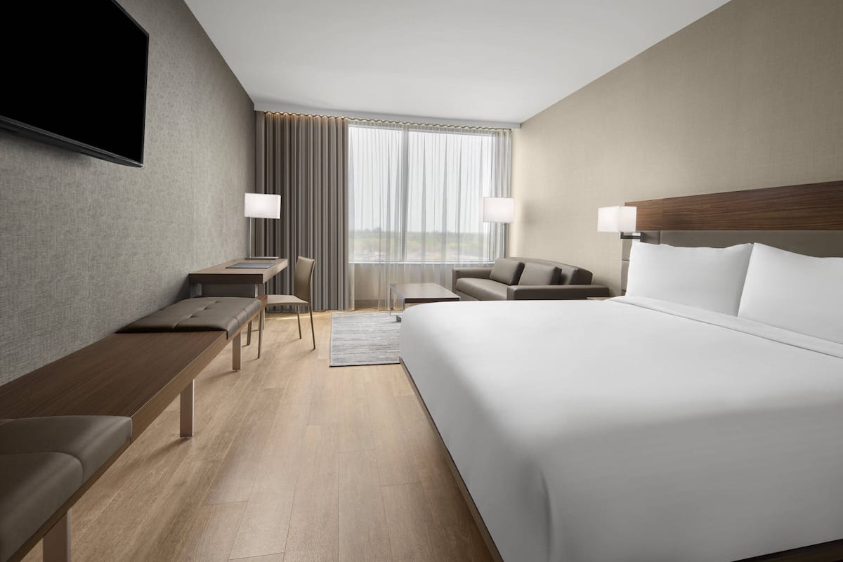 A modern hotel room with a large bed, a desk, a chair, a sofa, a TV, and a window with curtains.